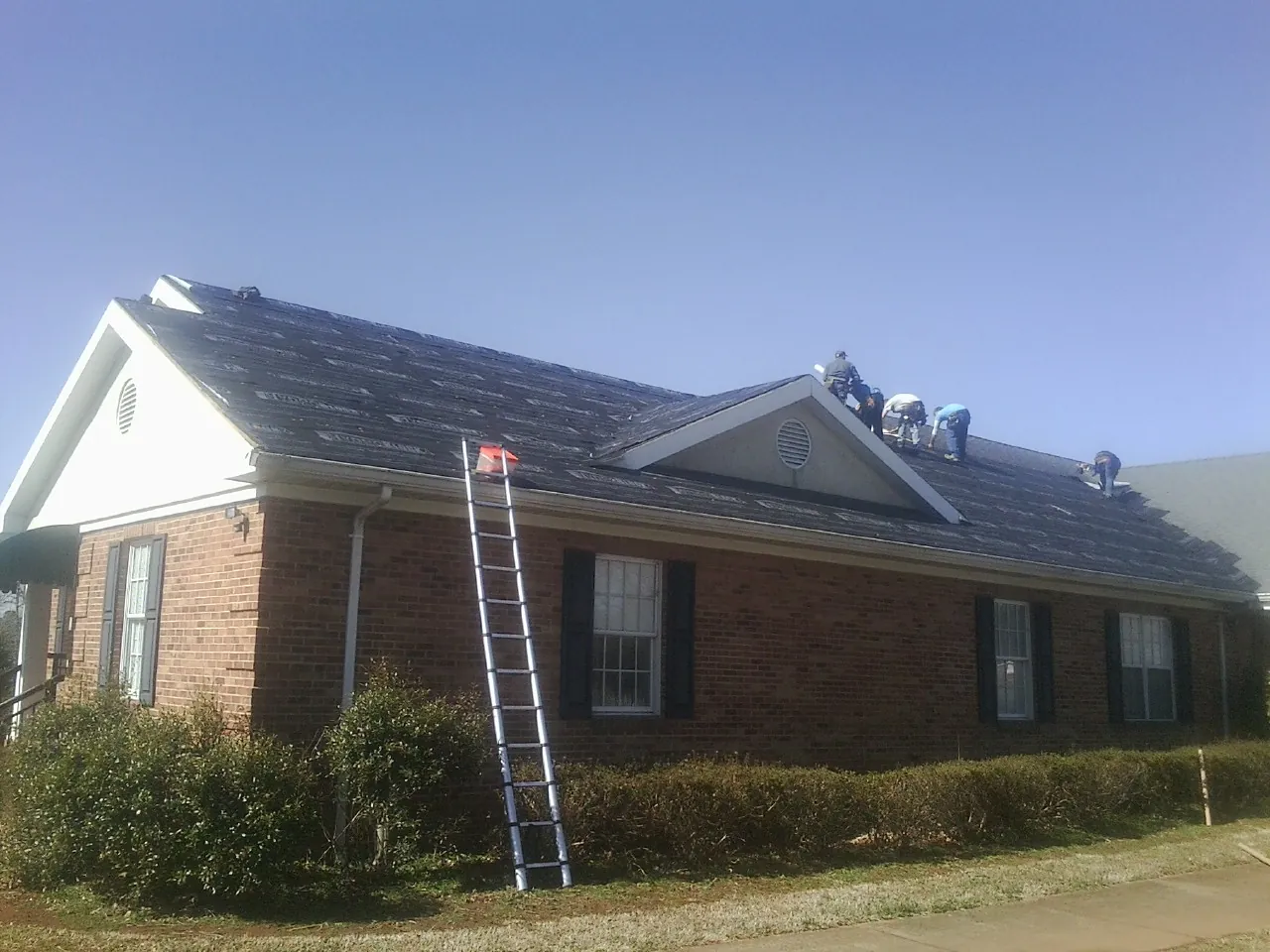 Crew tearing shingles off a brick ranch roof with a ladder set — Emergency Roof Repair work by our crew in Mineola