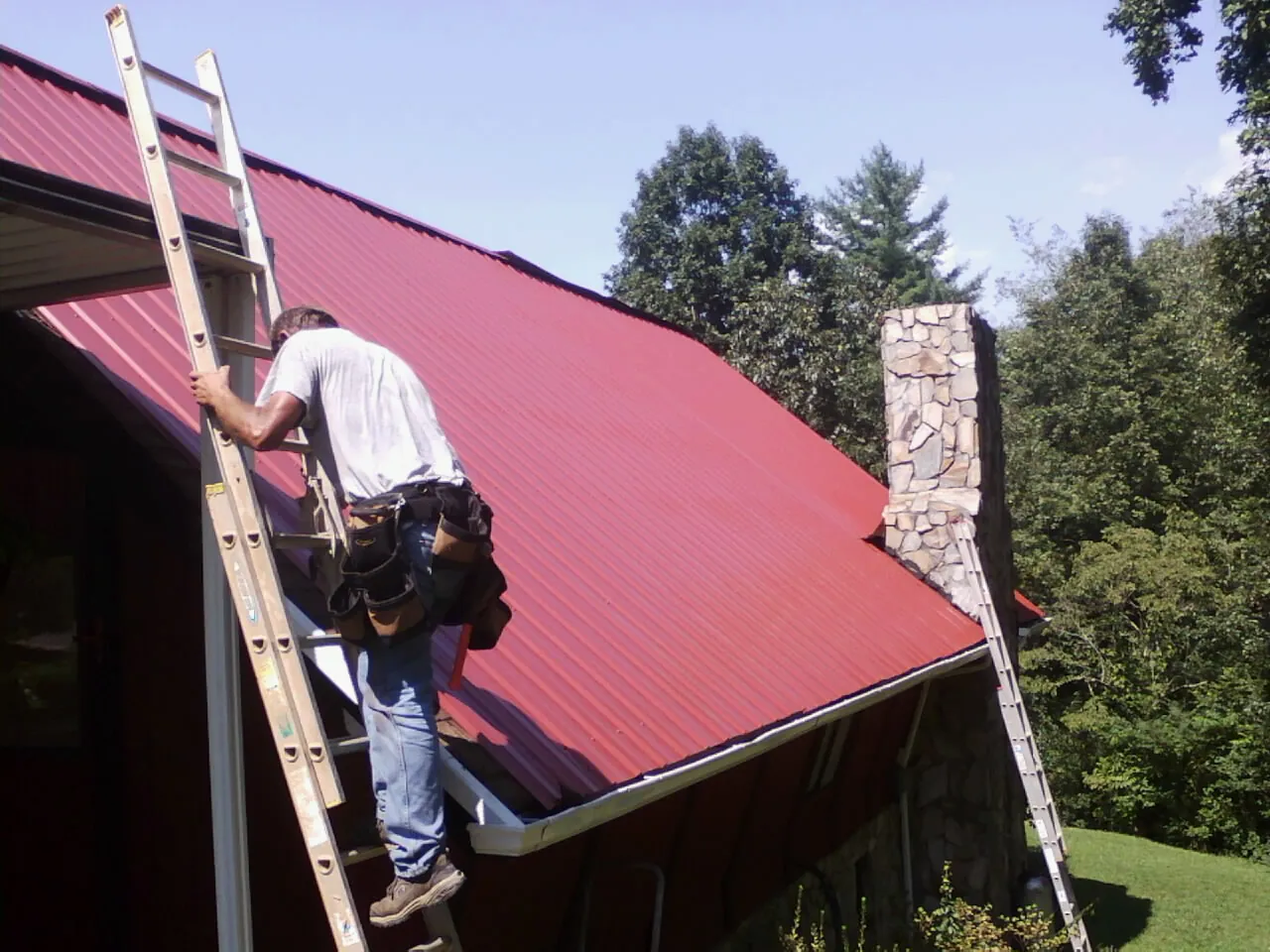Roofer on a ladder beside a red metal roof and stone chimney — Soffit & Fascia Repair by Roofing Brothers in Mineola